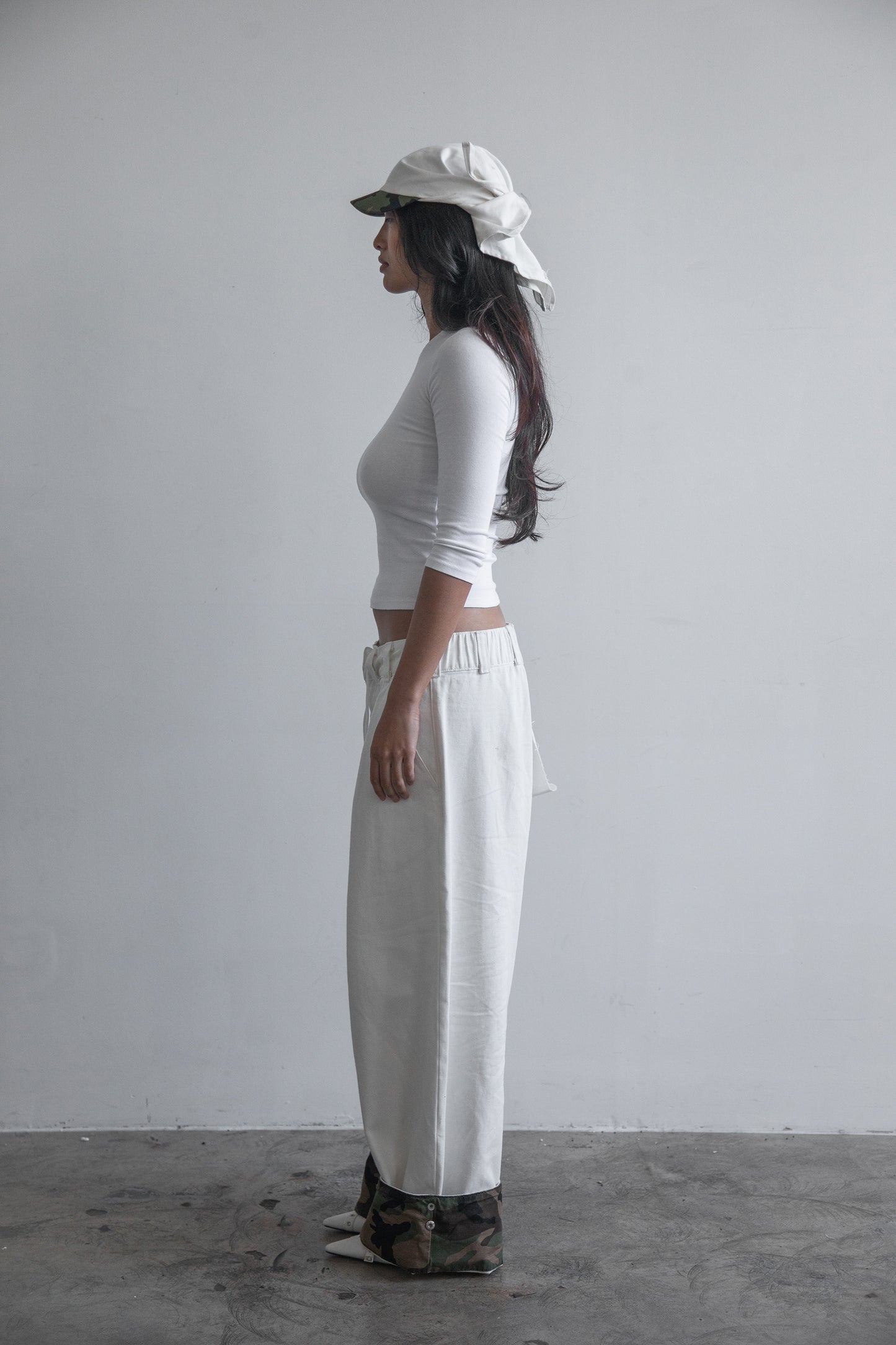 Side view of the DACY Studio Uniform Pants and Studio Tie Hat in off-white cotton drill, featuring adjustable camouflage hems, a short camouflage brim and oversized unisex fit.